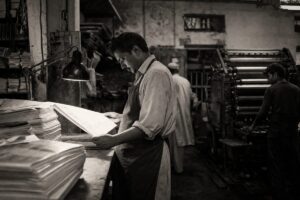 Newspaper printing press room in Pakistan, a pressman checking a freshly printed page under a work lamp (AI-generated image)