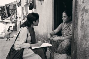 Researcher with a questionnaire sitting on a doorstep interviewing a resident, laundry lines overhead (AI-generated image)