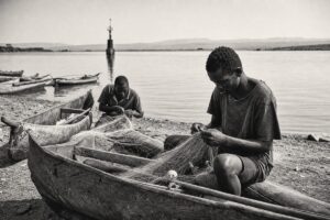 Fishermen mending nets beside wooden canoes on the shore of Lake Albert, an oil-survey marker in the distance (AI-generated image)