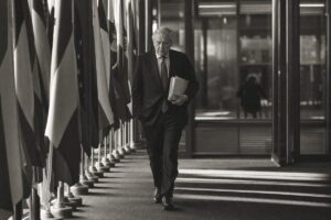 European institutional corridor lined with national flags, an official walking with briefing papers (AI-generated image)
