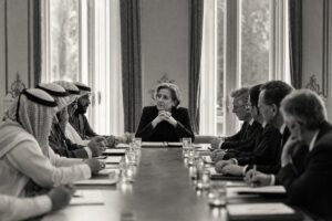 A mediator seated at the head of a negotiation table between two delegations, leaning forward to listen (AI-generated image)